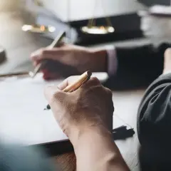 Dłonie piszące długopisami na dokumentach, w tle waga sprawiedliwości.